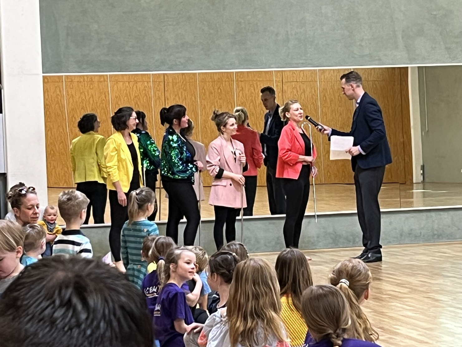 F&uuml;nf Frauen in bunten Outfits stehen auf einer B&uuml;hne, w&auml;hrend ein Mann sie interviewt. Zuschauer sitzen im Vordergrund.