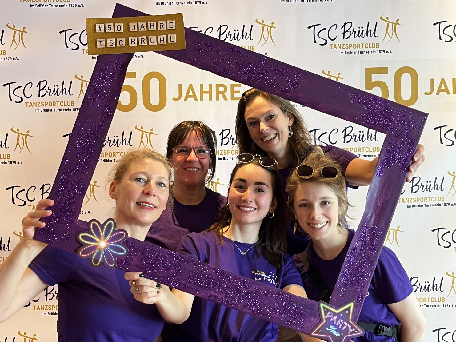 F&uuml;nf Frauen in lila T-Shirts posieren mit einem glitzernden Rahmen vor einem Banner zum 50-j&auml;hrigen Jubil&auml;um des TSC Br&uuml;hl.