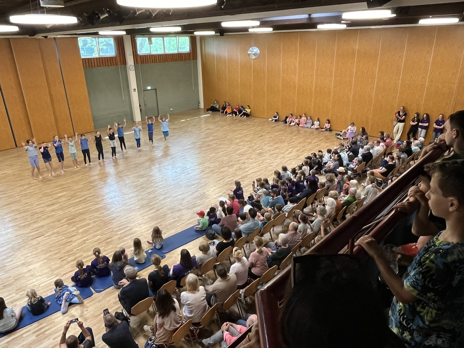 Tanzvorf&uuml;hrung mit mehreren T&auml;nzern auf einer Holzfl&auml;che, umgeben von einem Publikum auf Holzst&uuml;hlen.