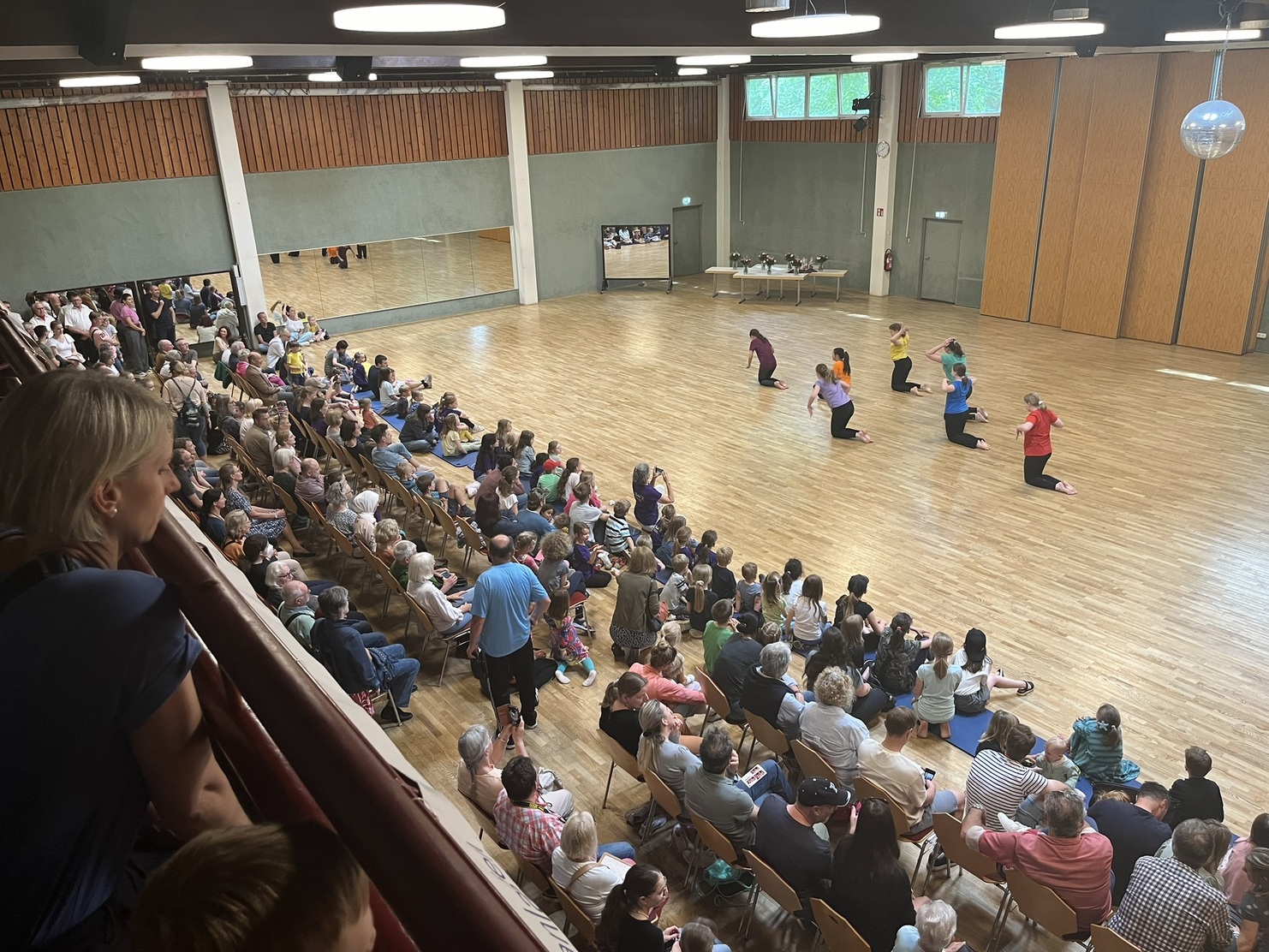 Tanzauff&uuml;hrung in einem gro&szlig;en Saal, umgeben von Zuschauern, mit T&auml;nzern in bunten Shirts auf dem Holzfu&szlig;boden.