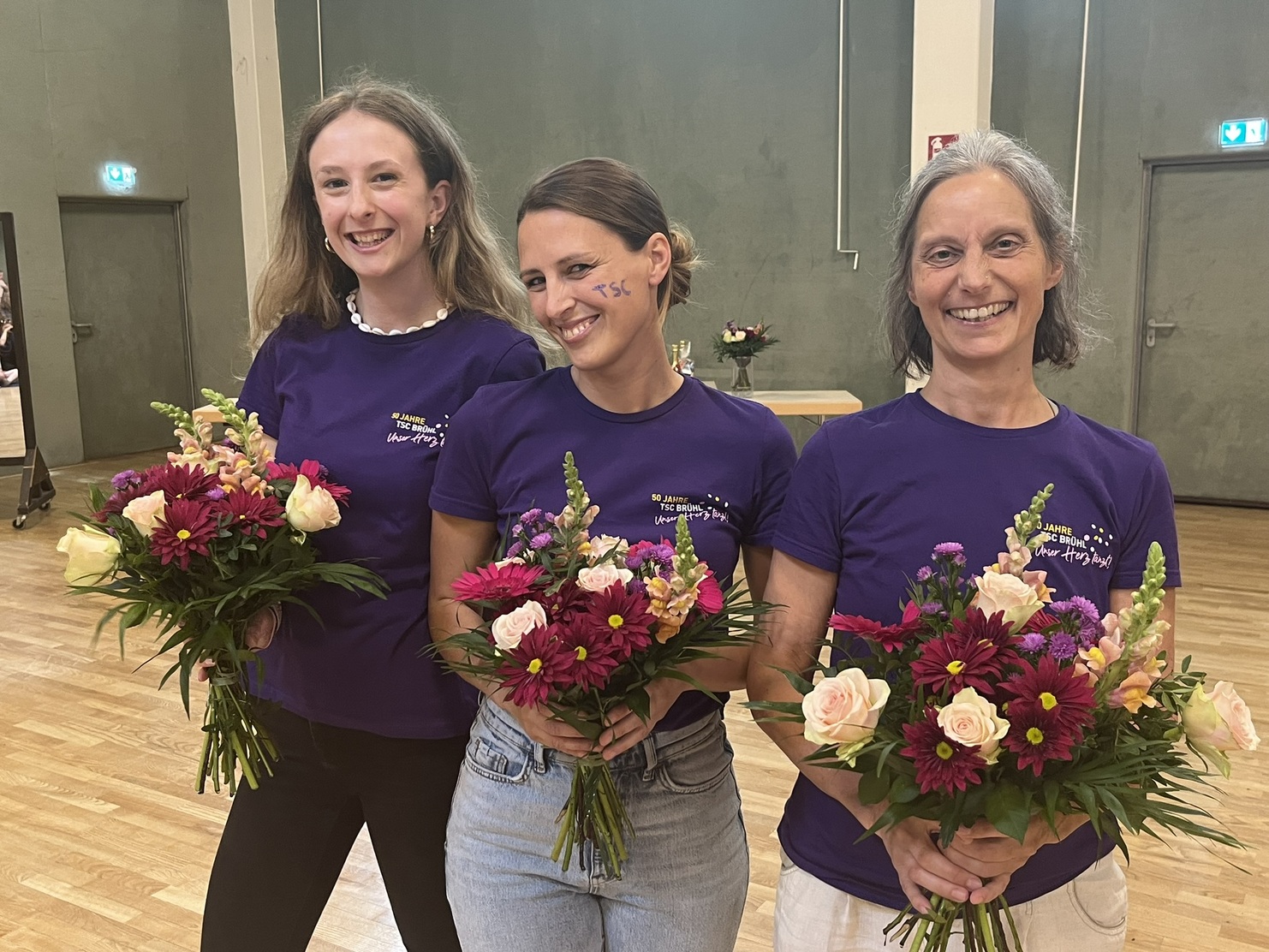 Drei Frauen in lila T-Shirts halten bunte Blumenstr&auml;u&szlig;e in einem gut beleuchteten Raum.