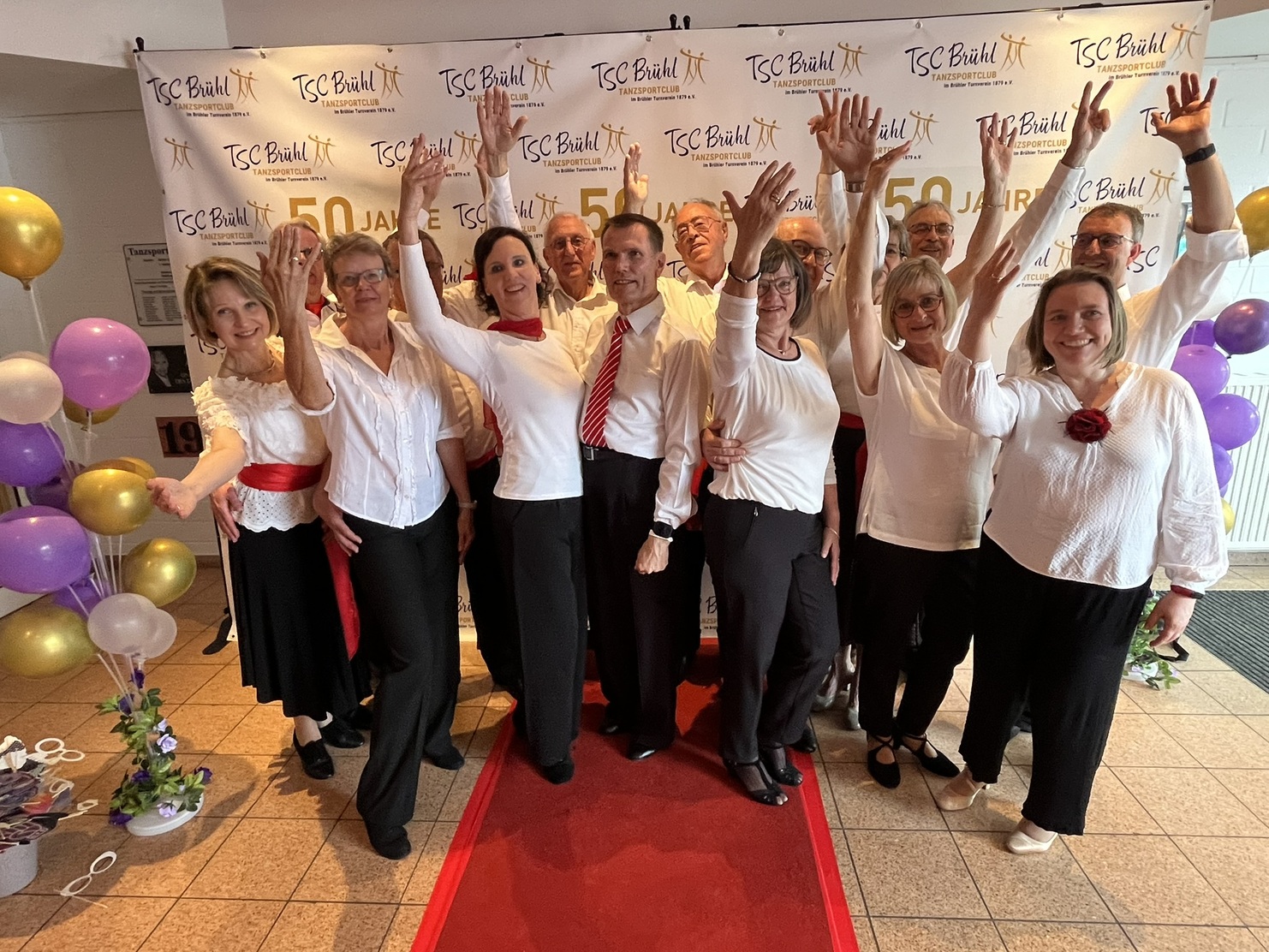 Gruppenselfie von T&auml;nzern in festlicher Kleidung, die vor einem Banner zum 50-j&auml;hrigen Jubil&auml;um posieren.