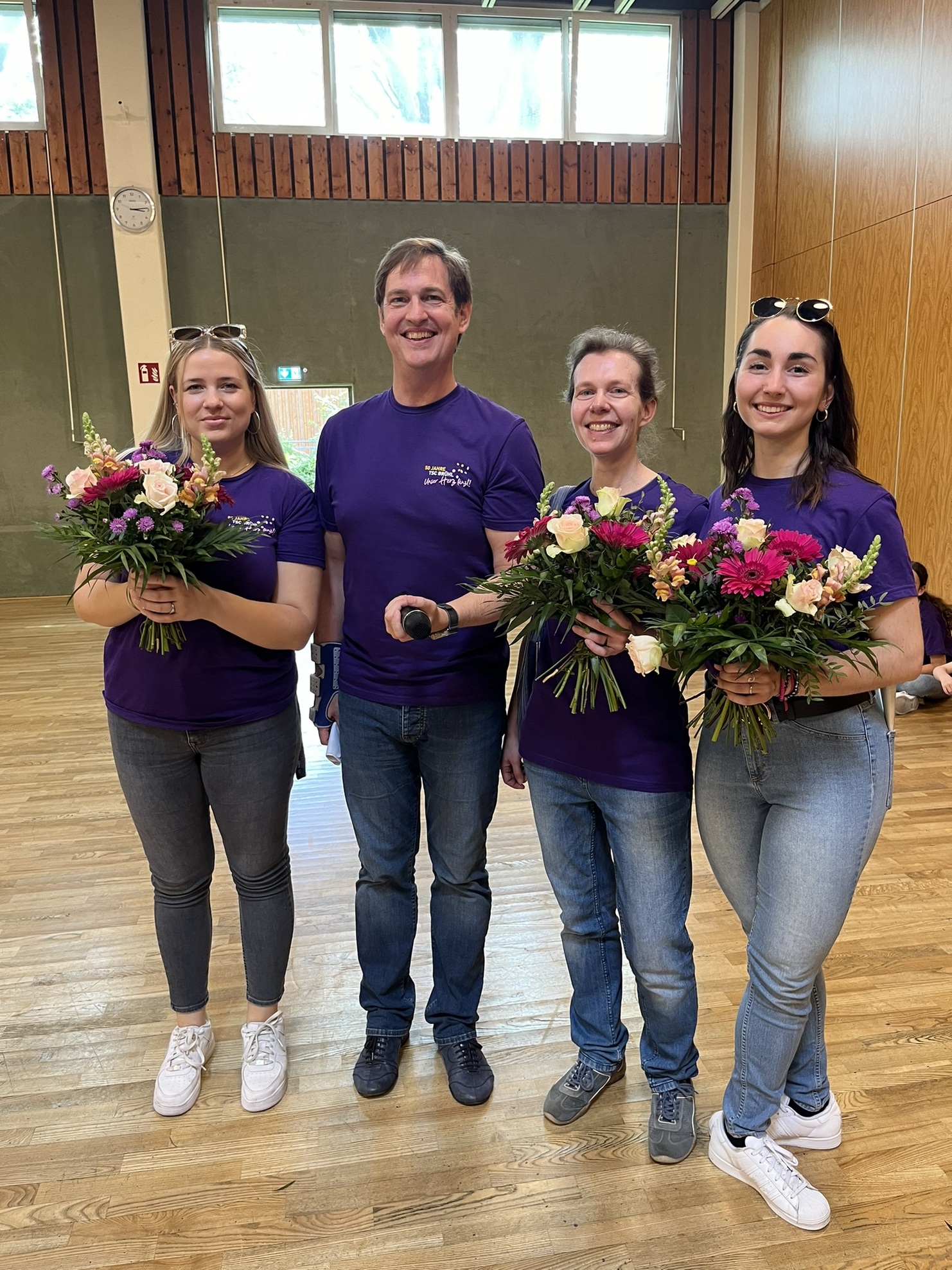 Vier Personen in lila T-Shirts halten Blumenstr&auml;u&szlig;e in einem Raum mit Holzboden. Im Hintergrund sind Fenster sichtbar.