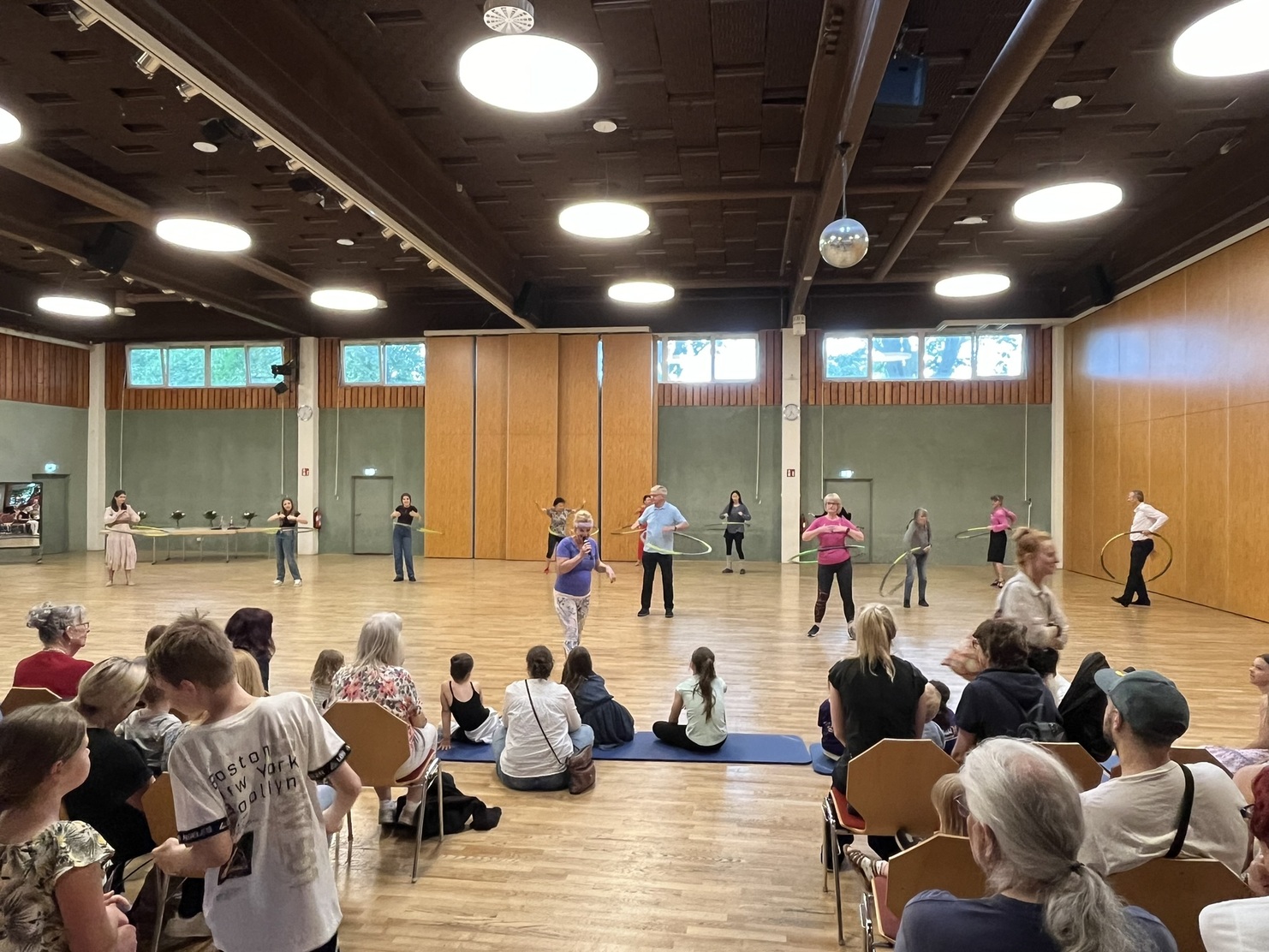 Gruppe von Menschen in einem Tanzsaal, die Hula-Hoop-Reifen in verschiedenen Farben nutzen, Publikum sitzt auf St&uuml;hlen.