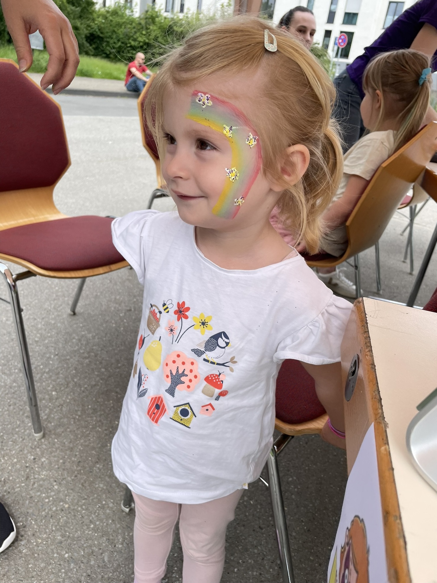 Kleines M&auml;dchen mit einem Regenbogen-Gesichtsmal in einem wei&szlig;en T-Shirt steht an einem Tisch, umgeben von St&uuml;hlen.