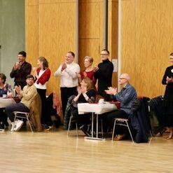 Eine Gruppe von Menschen applaudiert an Tischen in einem festlich dekorierten Saal bei einer Veranstaltung.