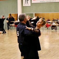 Tanzpaar in eleganter Kleidung f&uuml;hrt einen Turniertanz in einer Turnierhalle aus. Zuschauer im Hintergrund beobachten.