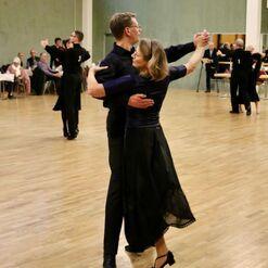 Tanzpaar in elegantem Outfit tanzt miteinander auf einer Holzfl&auml;che, Zuschauer im Hintergrund.