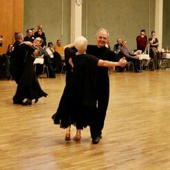 Tanzpaar in eleganter Abendkleidung tanzt auf einer Holzfl&auml;che, umgeben von Zuschauern.