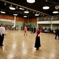 Tanzveranstaltung in einem gro&szlig;en Saal mit mehreren Paaren, die sich zur Musik bewegen, Tische und Zuschauer im Hintergrund.