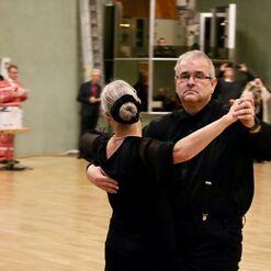 Tanzpaar in eleganter Kleidung beim Tanzen in einem Saal mit Holzfu&szlig;boden und Zuschauer im Hintergrund.