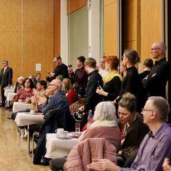 Personen sitzen und stehen applaudierend in einem Saal mit Holzw&auml;nden w&auml;hrend einer Veranstaltung. Tische sind sichtbar.