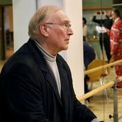 Ein &auml;lterer Mann mit Brille h&auml;lt Papier und einen Stift, w&auml;hrend er nachdenklich in einem hellen Raum steht.