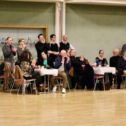 Gruppe von applaudierenden Menschen sitzt an Tischen in einem festlichen Raum mit Pokalen im Hintergrund.