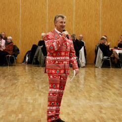Mann in rotem Weihnachtsanzug h&auml;lt Mikrophon und spricht vor Publikum in einem festlichen Saal.