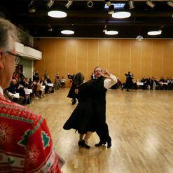 Tanzpaar in eleganter Kleidung f&uuml;hrt einen Walzer auf einer Tanzfl&auml;che, Zuschauer im Hintergrund beobachten aufmerksam.