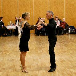 Tanzpaar in eleganter Kleidung, das auf einer Holzfl&auml;che eng miteinander tanzt, w&auml;hrend Zuschauer im Hintergrund zuschauen.