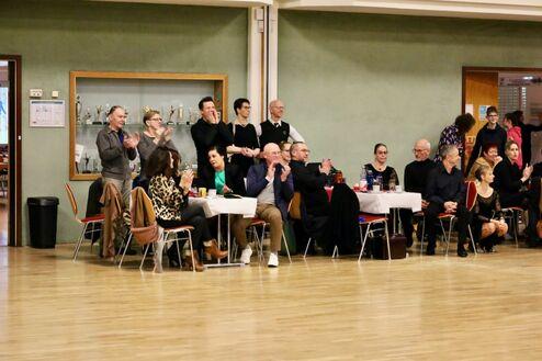 Gruppe von applaudierenden Menschen sitzt an Tischen in einem festlichen Raum mit Pokalen im Hintergrund.