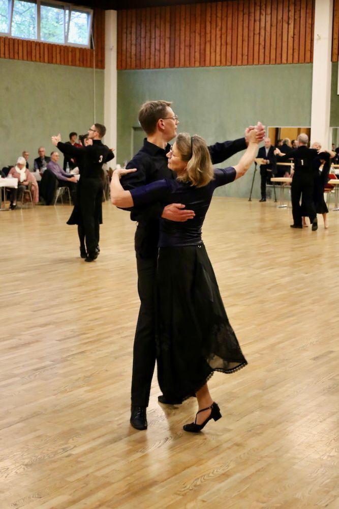 Tanzpaar in elegantem Outfit tanzt miteinander auf einer Holzfl&auml;che, Zuschauer im Hintergrund.