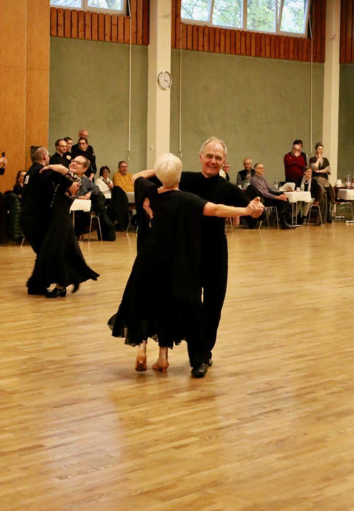 Tanzpaar in eleganter Abendkleidung tanzt auf einer Holzfl&auml;che, umgeben von Zuschauern.