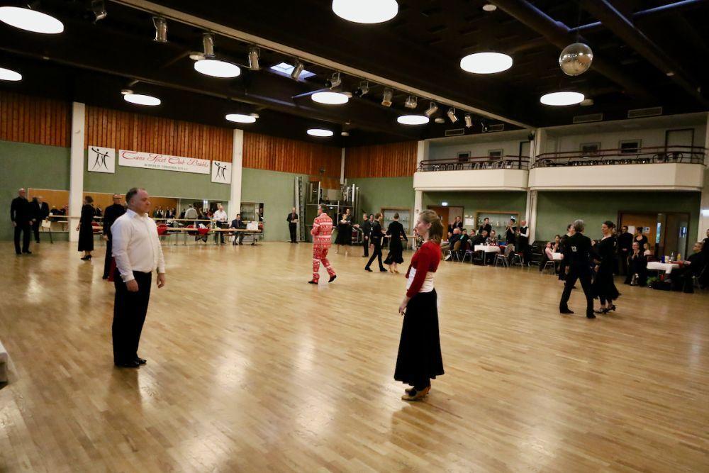 Tanzveranstaltung in einem gro&szlig;en Saal mit mehreren Paaren, die sich zur Musik bewegen, Tische und Zuschauer im Hintergrund.