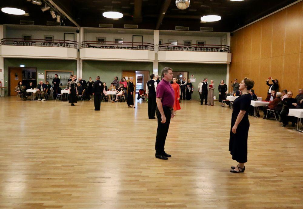 Tanzpaar steht sich auf einer Tanzfl&auml;che gegen&uuml;ber, Zuschauer sitzen an Tischen im Hintergrund.