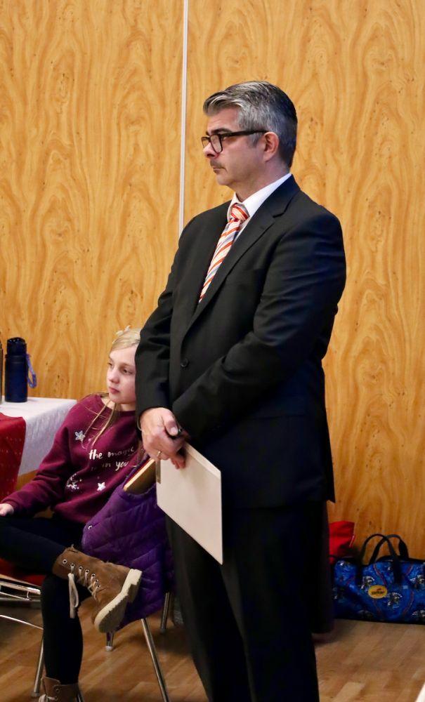 Ein Mann im Anzug steht neben einem Sitzplatz, w&auml;hrend ein M&auml;dchen in einem Pullover und einer Weste sitzt.