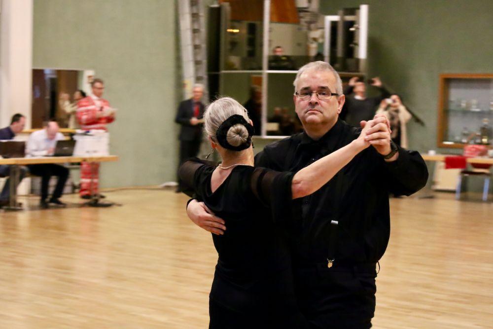 Tanzpaar in eleganter Kleidung beim Tanzen in einem Saal mit Holzfu&szlig;boden und Zuschauer im Hintergrund.