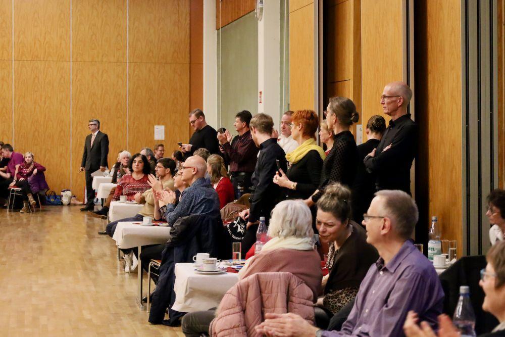 Personen sitzen und stehen applaudierend in einem Saal mit Holzw&auml;nden w&auml;hrend einer Veranstaltung. Tische sind sichtbar.