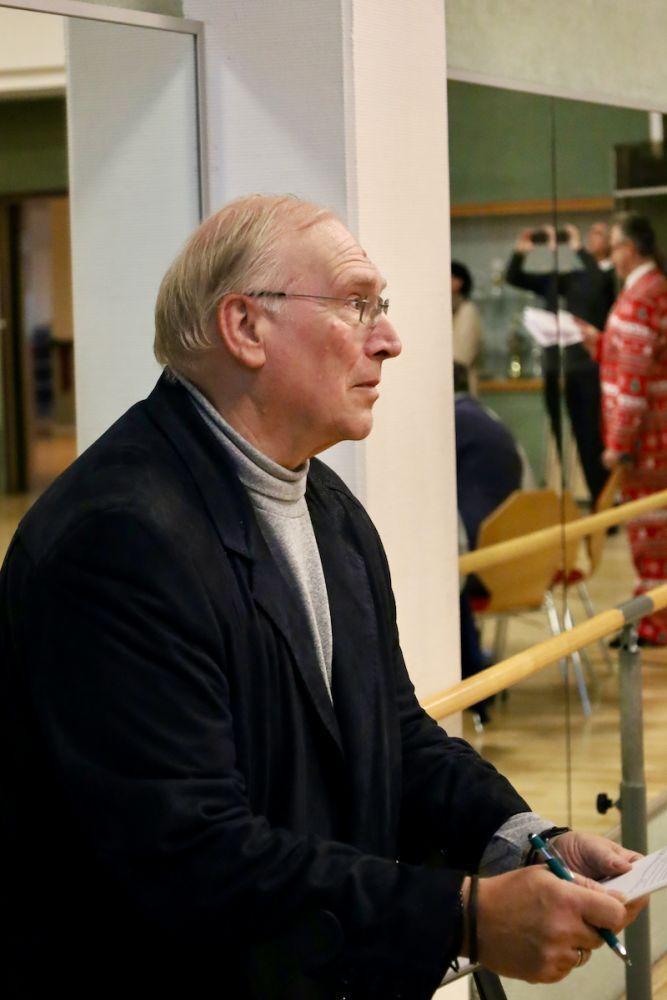 Ein &auml;lterer Mann mit Brille h&auml;lt Papier und einen Stift, w&auml;hrend er nachdenklich in einem hellen Raum steht.