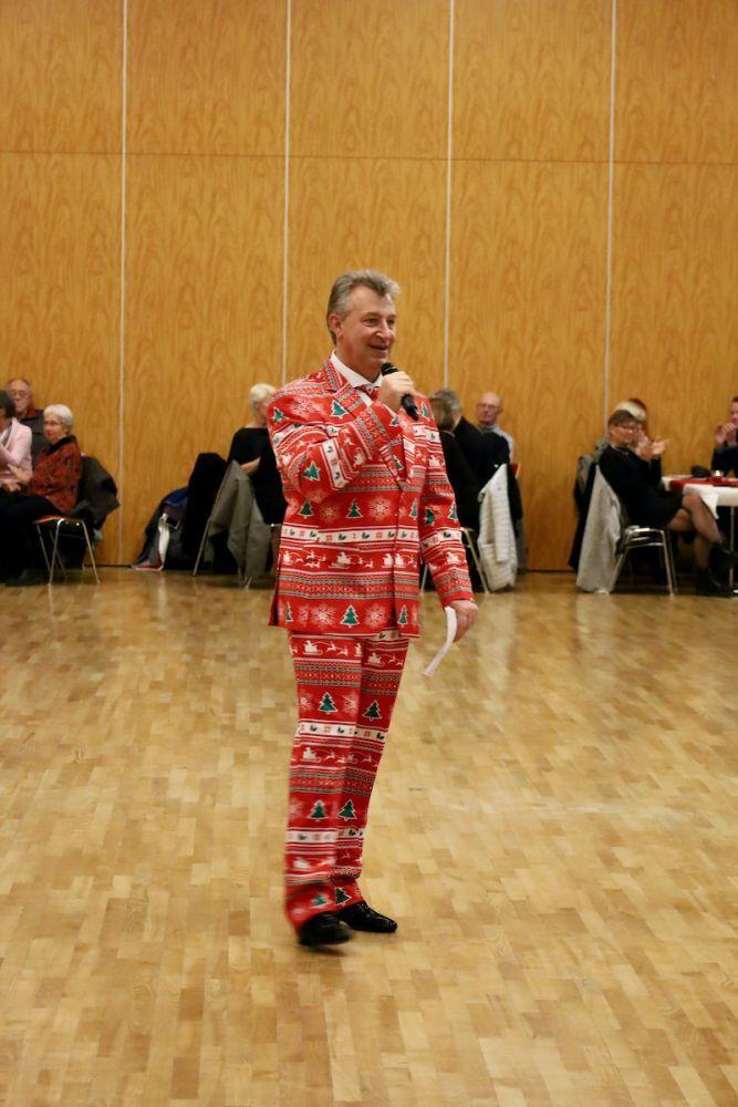 Mann in rotem Weihnachtsanzug h&auml;lt Mikrophon und spricht vor Publikum in einem festlichen Saal.