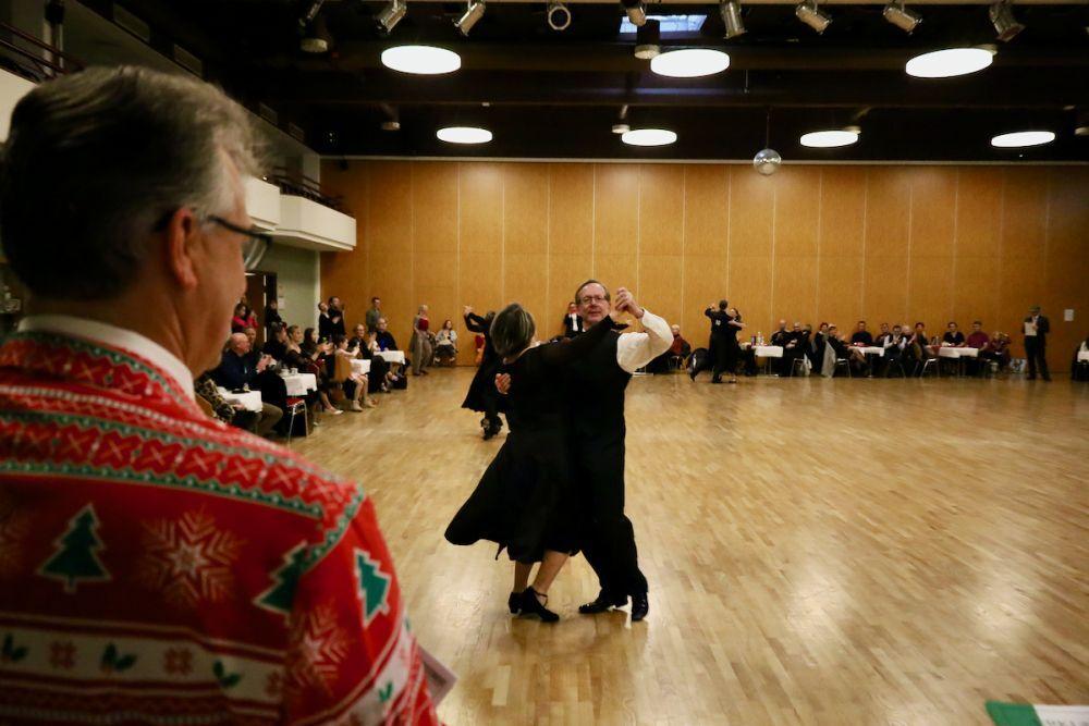 Tanzpaar in eleganter Kleidung f&uuml;hrt einen Walzer auf einer Tanzfl&auml;che, Zuschauer im Hintergrund beobachten aufmerksam.