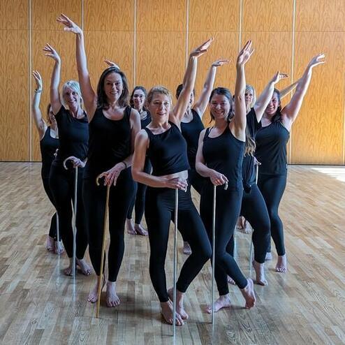 Gruppe von Frauen in schwarzer Kleidung, die mit Stäben tanzen, auf einem hellen Parkettboden.