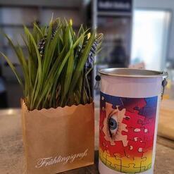 Topf mit grünen Pflanzen in einer Papiertüte neben einem bunten Eimer mit einem Puzzle-Motiv und einer Augen-Illustration.