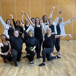 Gruppe von zw&ouml;lf Frauen in schwarzer Kleidung posiert fr&ouml;hlich in einem Raum mit Holzboden.