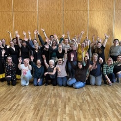 Gruppensnapshot von 40 Personen in einem Raum, die fr&ouml;hlich die H&auml;nde heben. Ein paar tragen farbenfrohe Kleidung.