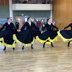 Vier T&auml;nzerinnen in schwarzen Kleidern mit gelben R&uuml;schen tanzen synchron in einem gro&szlig;en Raum.