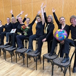 Gruppe von Frauen in schwarzer Kleidung sitzt auf St&uuml;hlen, hebt die H&auml;nde und h&auml;lt Blumen sowie eine farbige Ballonfigur.