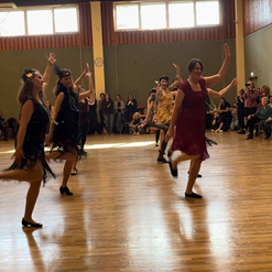 Tanzende Frauen in festlichen Kleidern zeigen synchronen Tanz auf einer Holzb&uuml;hne vor Publikum.