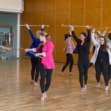 Gruppe von Frauen in Leggings und Oberteilen, die mit St&ouml;cken in einem Tanzraum mit Holzboden tanzen.