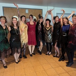 Zehn Frauen in stilvollen 1920er-Jahre-Kleidern posieren fr&ouml;hlich in einer Innenraum-Umgebung.