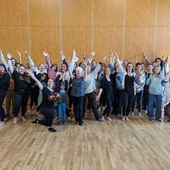 Gruppenfoto von 30 Personen, die in einem Raum mit Holzfußboden stehen und jubelnd die Hände heben.