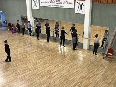 Gruppe von Tänzern an einer Ballettstange, reflektiert in Spiegeln, in einem Tanzstudio mit Holzfußboden.