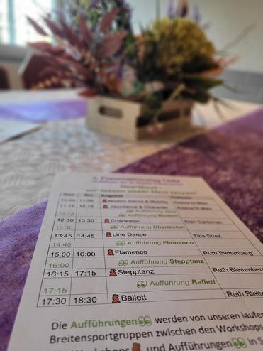 Tisch mit einem Programmheft für Tanzaufführungen und einer blühenden Dekoration im Hintergrund.