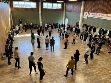 Gruppe von T&auml;nzern in einem Saal, die sich in einem gro&szlig;en Kreis umarmen und miteinander tanzen.