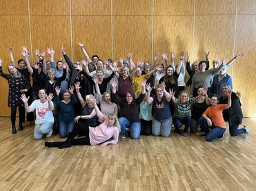 Gruppenbild mit vielen Menschen, die fr&ouml;hlich die H&auml;nde heben, auf Holzfu&szlig;boden vor einer Wand aus Holzpaneelen.