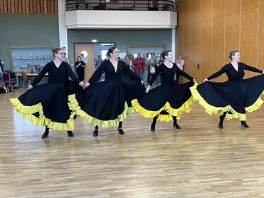 Vier T&auml;nzerinnen in schwarzen Kleidern mit gelben R&uuml;schen tanzen synchron in einem gro&szlig;en Raum.