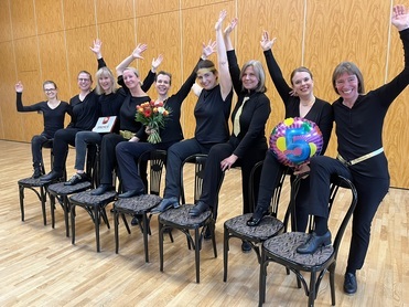 Gruppe von Frauen in schwarzer Kleidung sitzt auf St&uuml;hlen, hebt die H&auml;nde und h&auml;lt Blumen sowie eine farbige Ballonfigur.