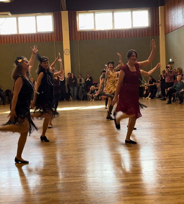Tanzende Frauen in festlichen Kleidern zeigen synchronen Tanz auf einer Holzb&uuml;hne vor Publikum.
