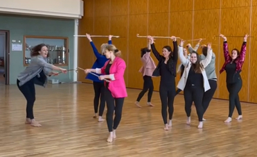 Gruppe von Frauen in Leggings und Oberteilen, die mit St&ouml;cken in einem Tanzraum mit Holzboden tanzen.
